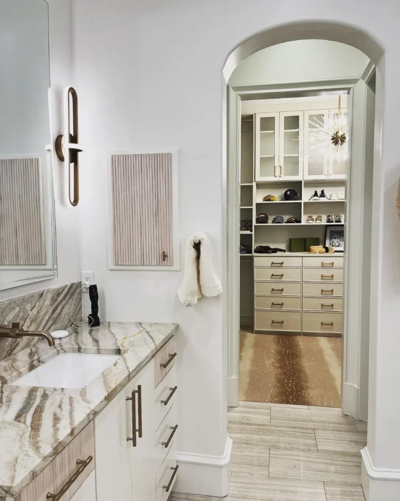 Luxury bathroom vanity with marble countertops leading into a custom walk-in closet with built-in cabinetry and shelving.