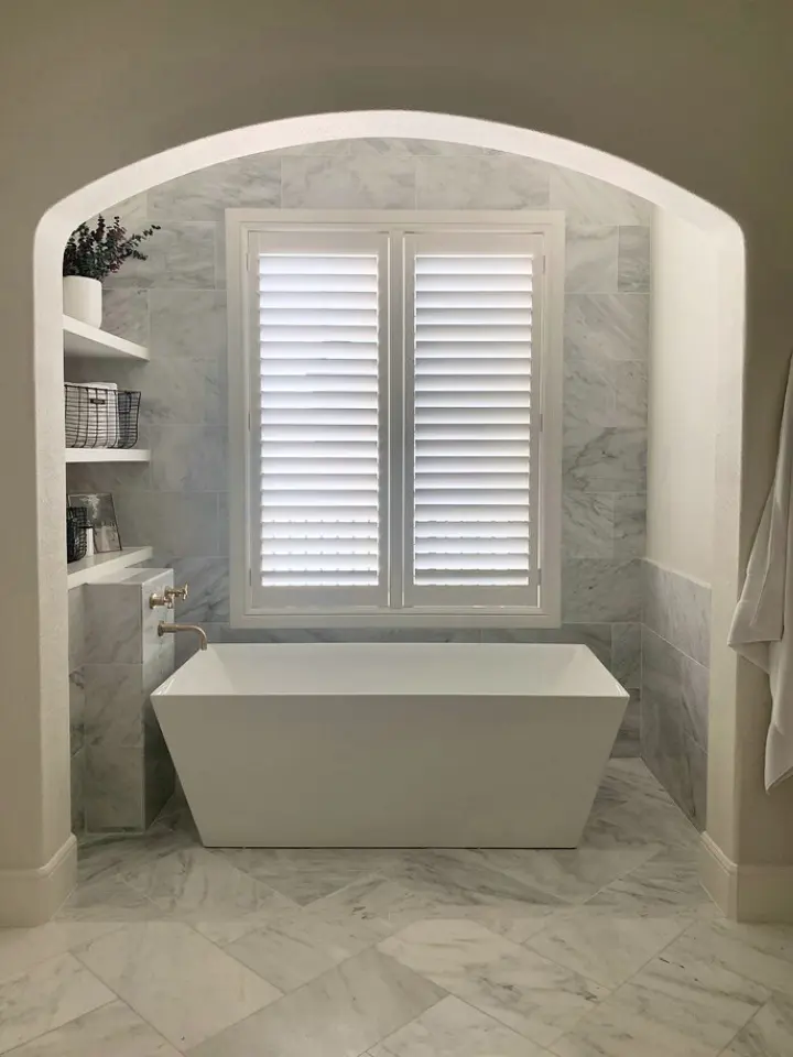 Freestanding white bathtub beneath shuttered windows in a marble bathroom alcove with arched entryway.