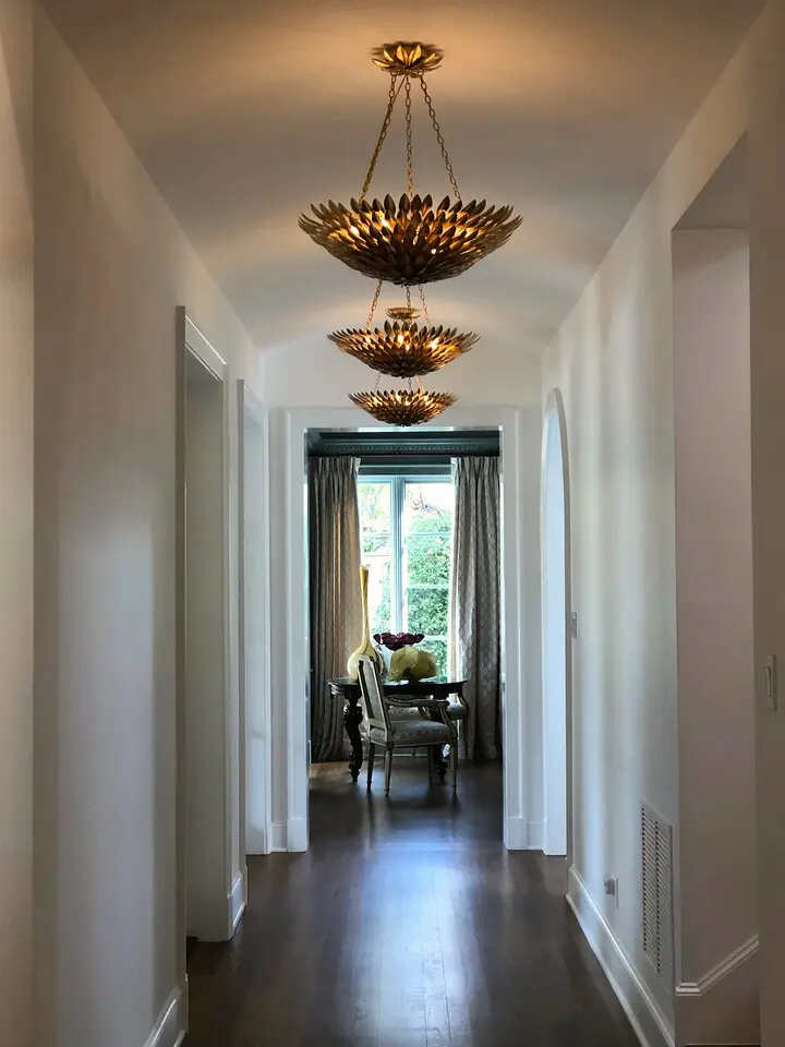 Long hallway with sculptural pendant chandeliers, dark wood flooring, and natural light from a windowed sitting area.