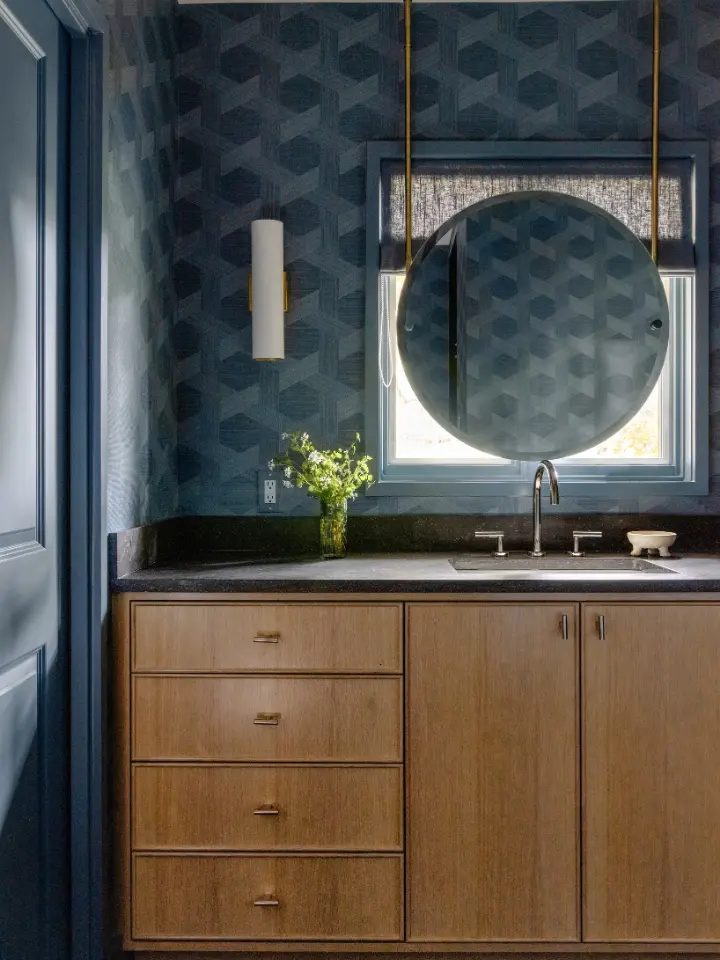 Modern bathroom vanity with wood cabinetry, dark geometric wallpaper, round mirror, and black stone countertop.