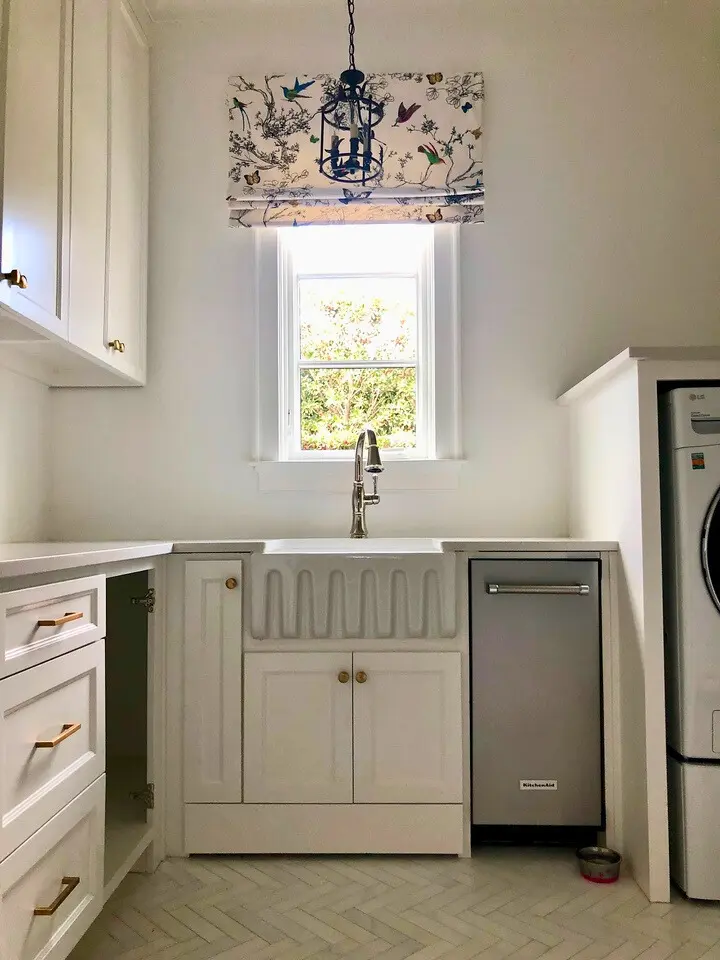 Bright laundry room with farmhouse sink, white cabinetry, patterned window shade, and built-in appliances.