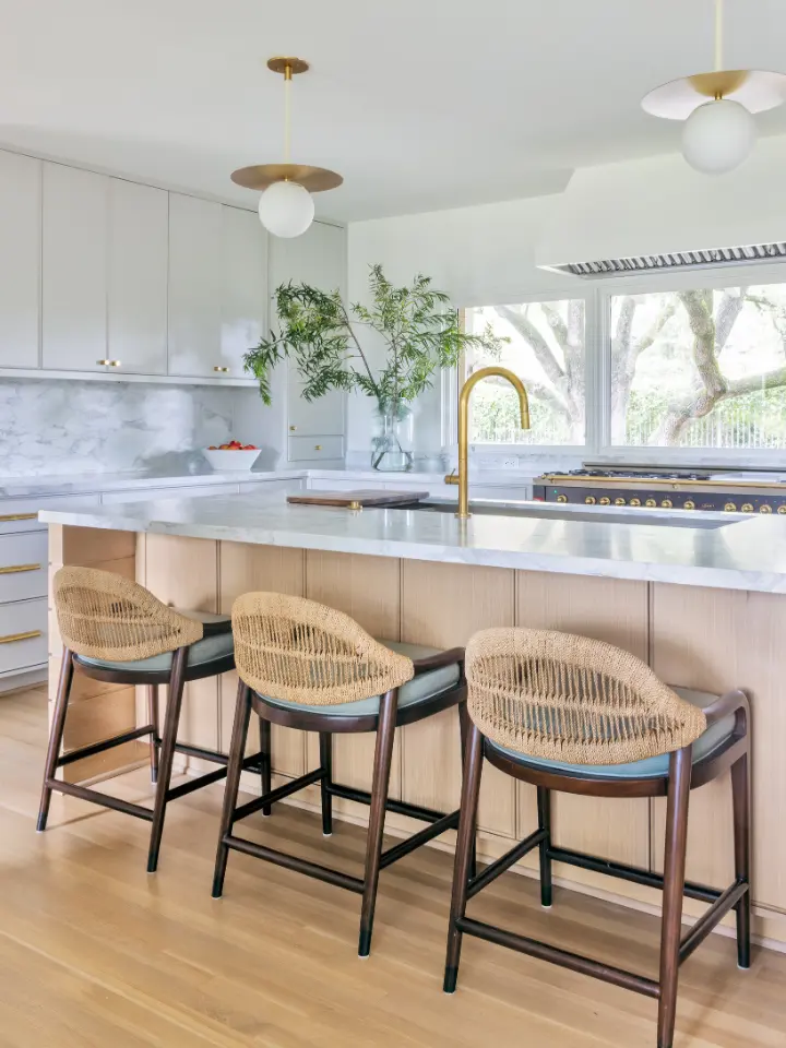 Bright modern kitchen with waterfall island, woven bar stools, brass faucet, and large windows.