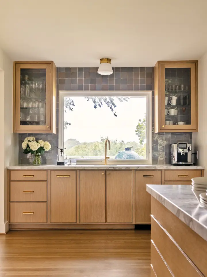 Modern kitchen with natural wood cabinetry, brass hardware, tiled backsplash, and large picture window above the sink.
