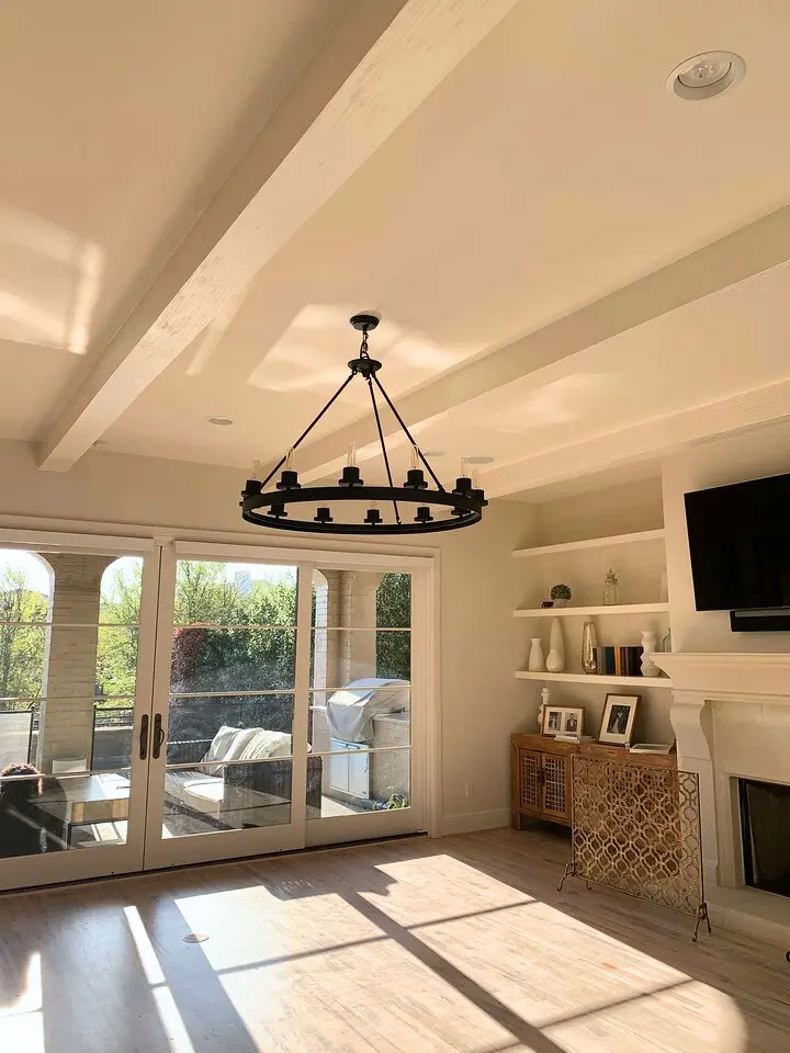 Bright living room with exposed ceiling beams, circular chandelier, fireplace, and French doors leading to an outdoor patio.