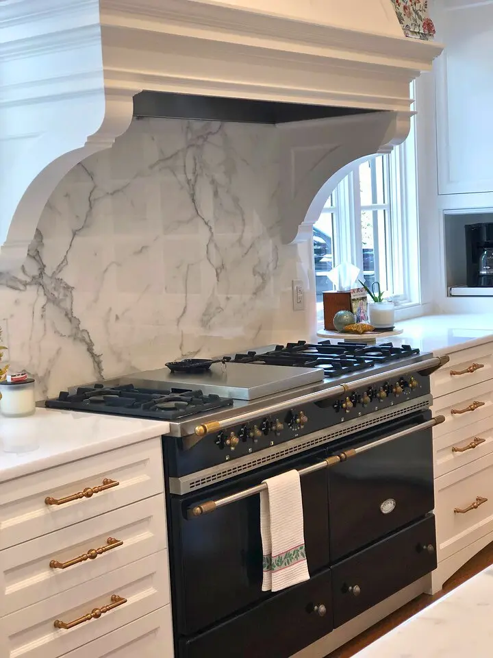 Luxury kitchen with marble backsplash, custom range hood, and black gas range with brass accents.