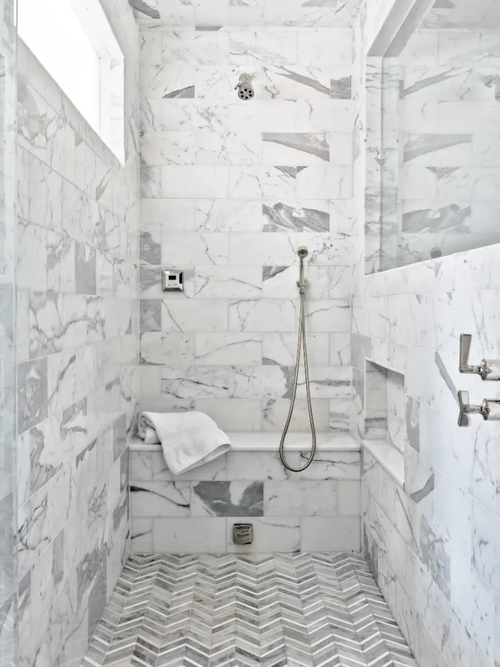 Luxury marble walk-in shower with built-in bench, handheld shower fixture, and chevron tile flooring.