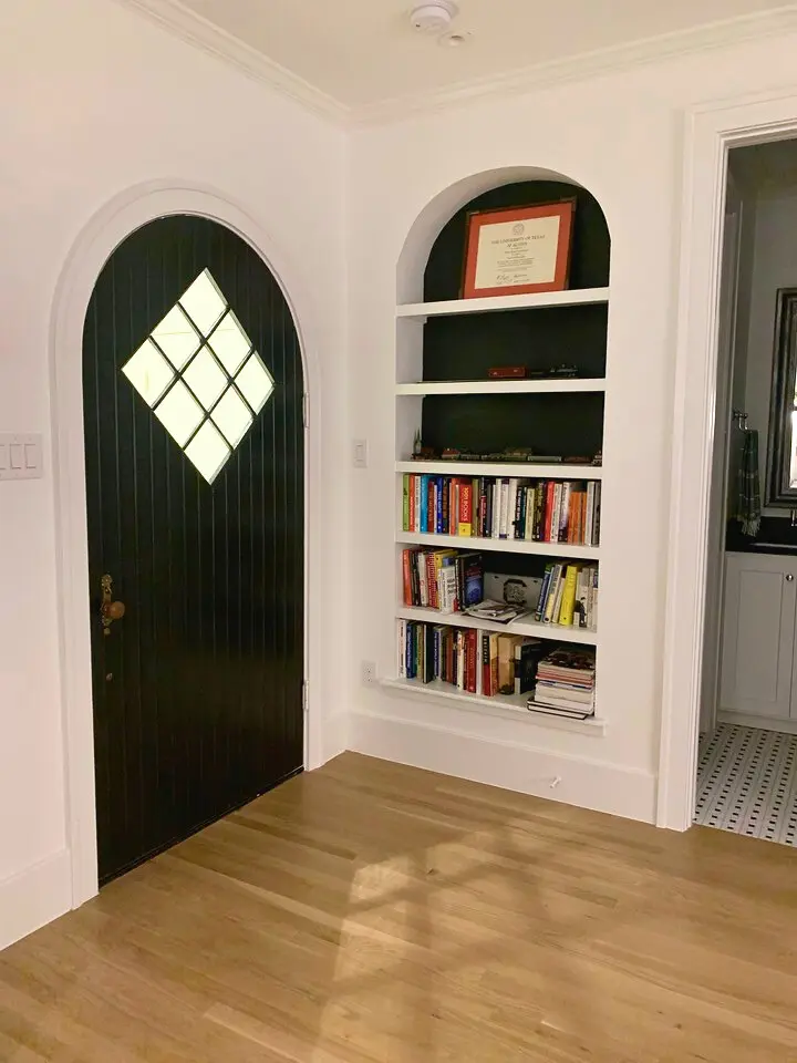 Arched black entry door with diamond window detail beside a built-in bookshelf in a bright modern interior.