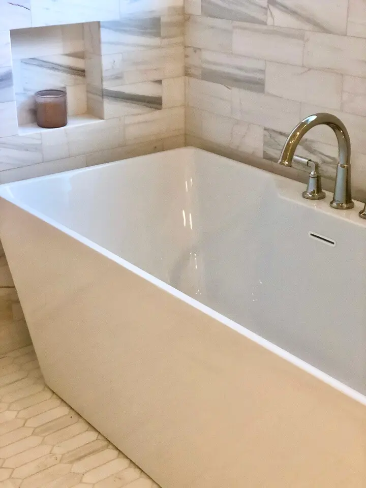 Modern freestanding bathtub with brass fixtures, marble tile walls, and hexagon tile flooring.