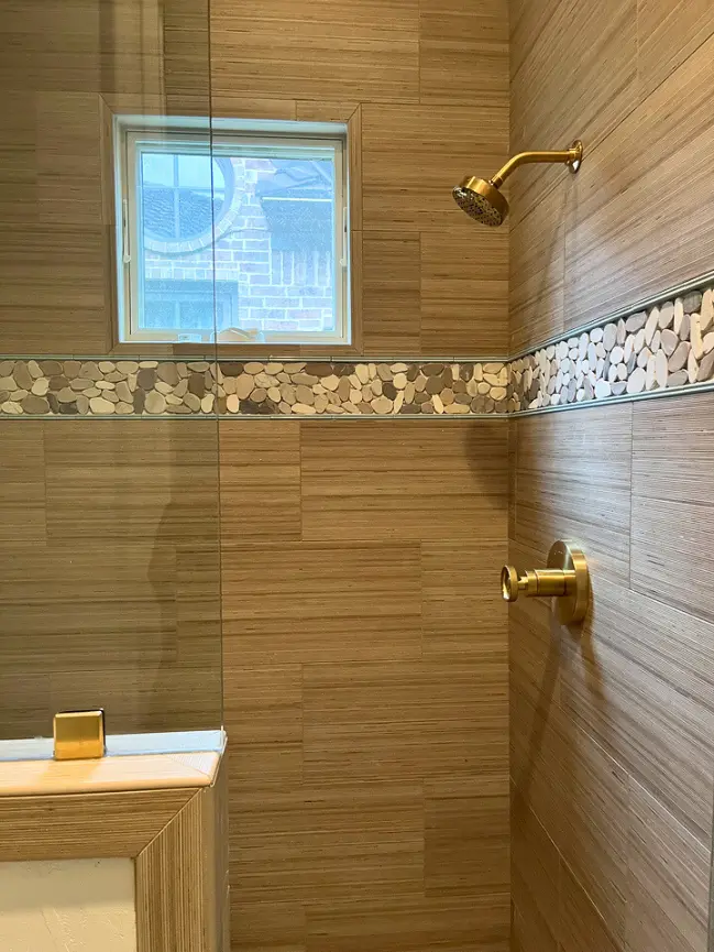 Modern walk-in shower with textured wood-look tile walls, pebble accent border, and brass shower fixtures.