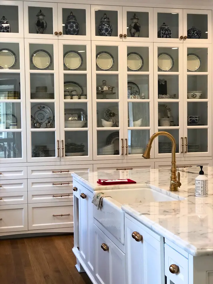 Luxury white kitchen with glass-front cabinetry, marble countertops, farmhouse sink, and brass faucet fixtures.