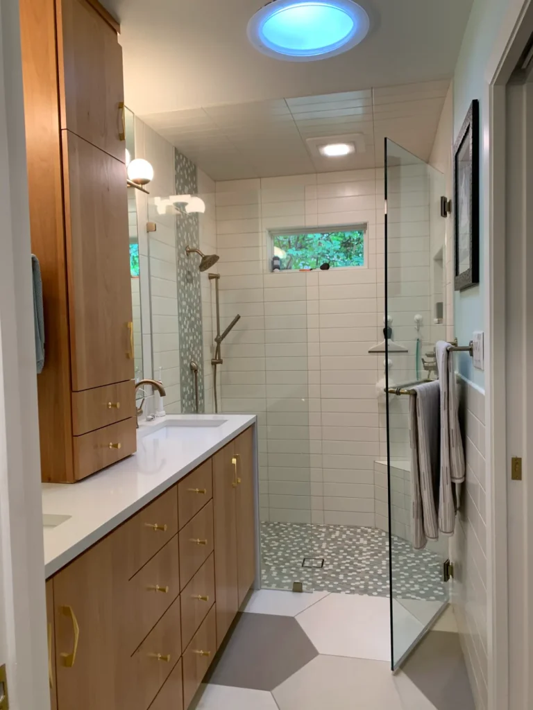 Modern bathroom remodel with wood vanity, quartz countertop, frameless glass shower, white tile walls, and mosaic shower flooring.