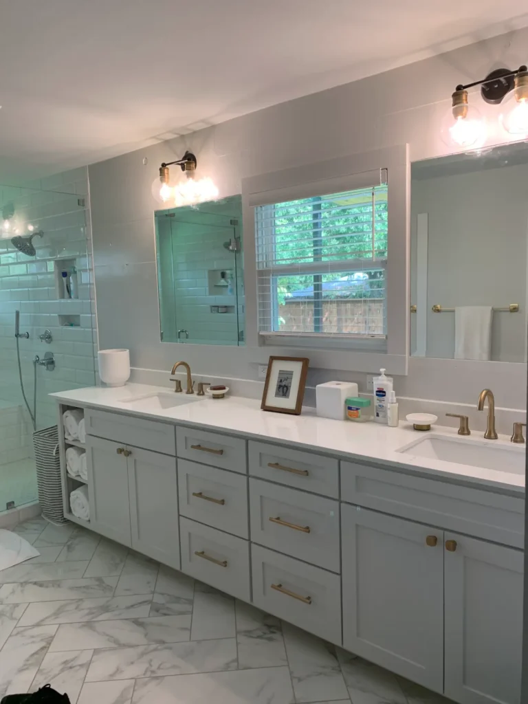 Modern bathroom remodel with double vanity, gray shaker cabinets, quartz countertop, brass fixtures, large mirrors, and marble tile flooring.