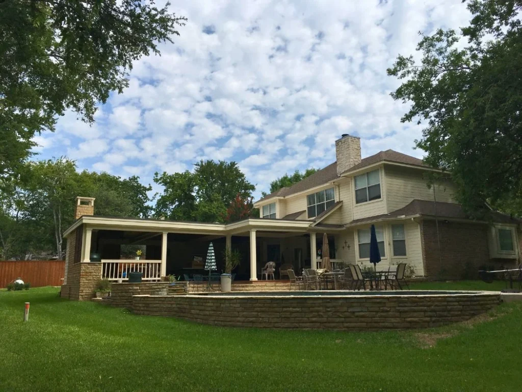 Large backyard outdoor living space with covered patio, stone fireplace, seating area, and expansive lawn behind a custom home