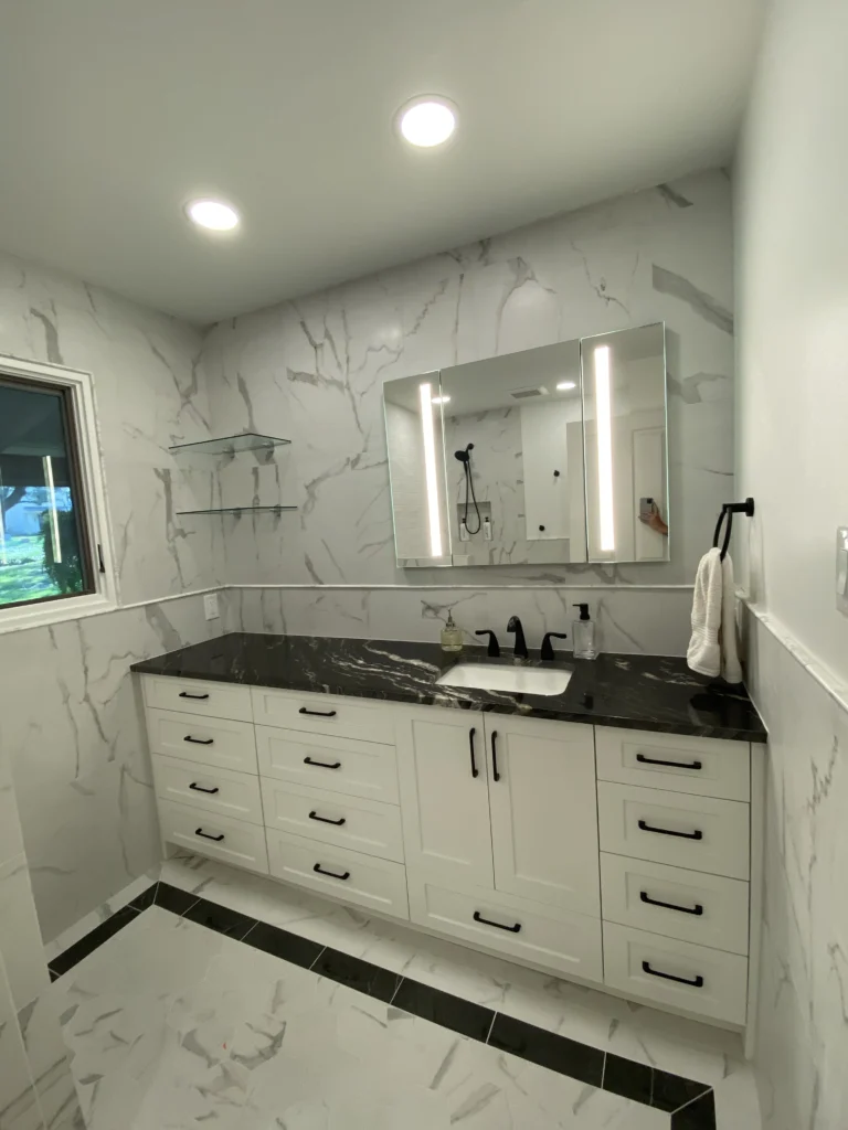 Modern bathroom vanity with marble walls, black countertops, illuminated mirror, and custom white cabinetry